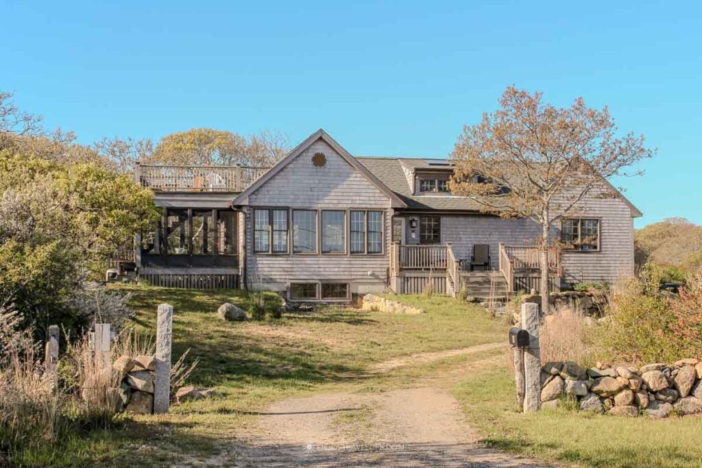A shingled rental house with a screened porch and wraparound deck set back on a grassy lot behind a stone wall and dirt path in Aquinnah