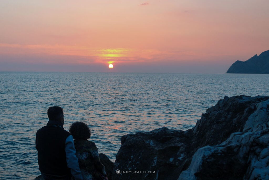 A couple silhouetted on the harbor rocks in Vernazza watching the sunset over the Tyrrhenian Sea in Cinque Terre