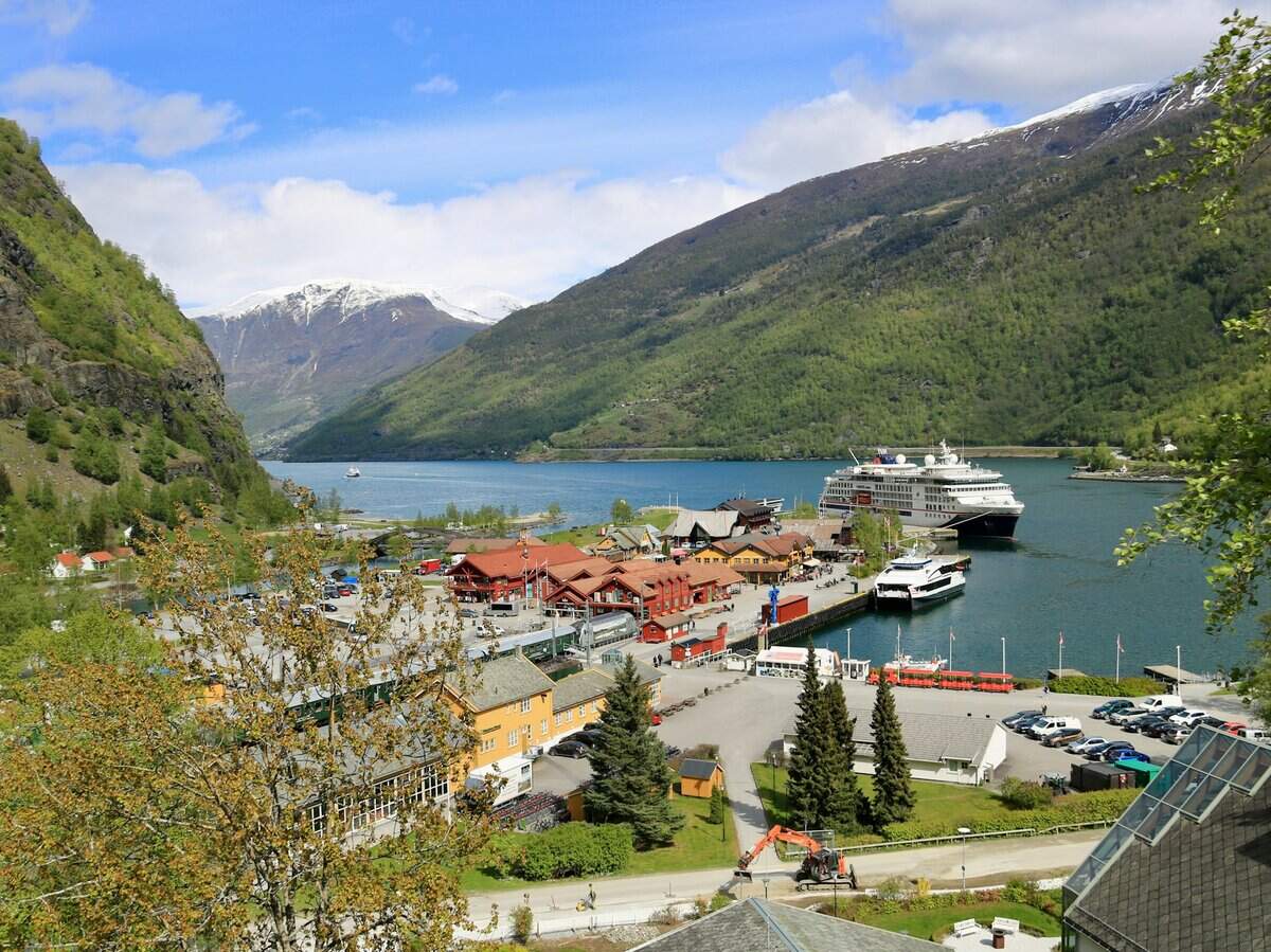 Cruise ship docked at the Flam pier with colorful village buildings and snow-capped mountains in the background.