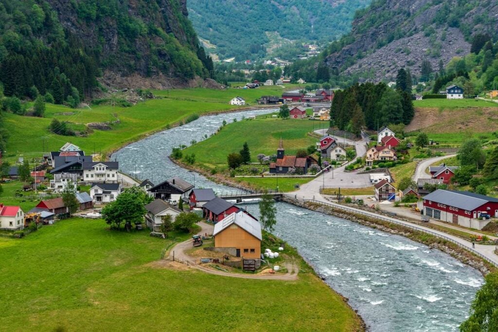 Colorful houses line both sides of the Flam River in the village of Flam, Norway, with green fields and mountains surrounding the valley.