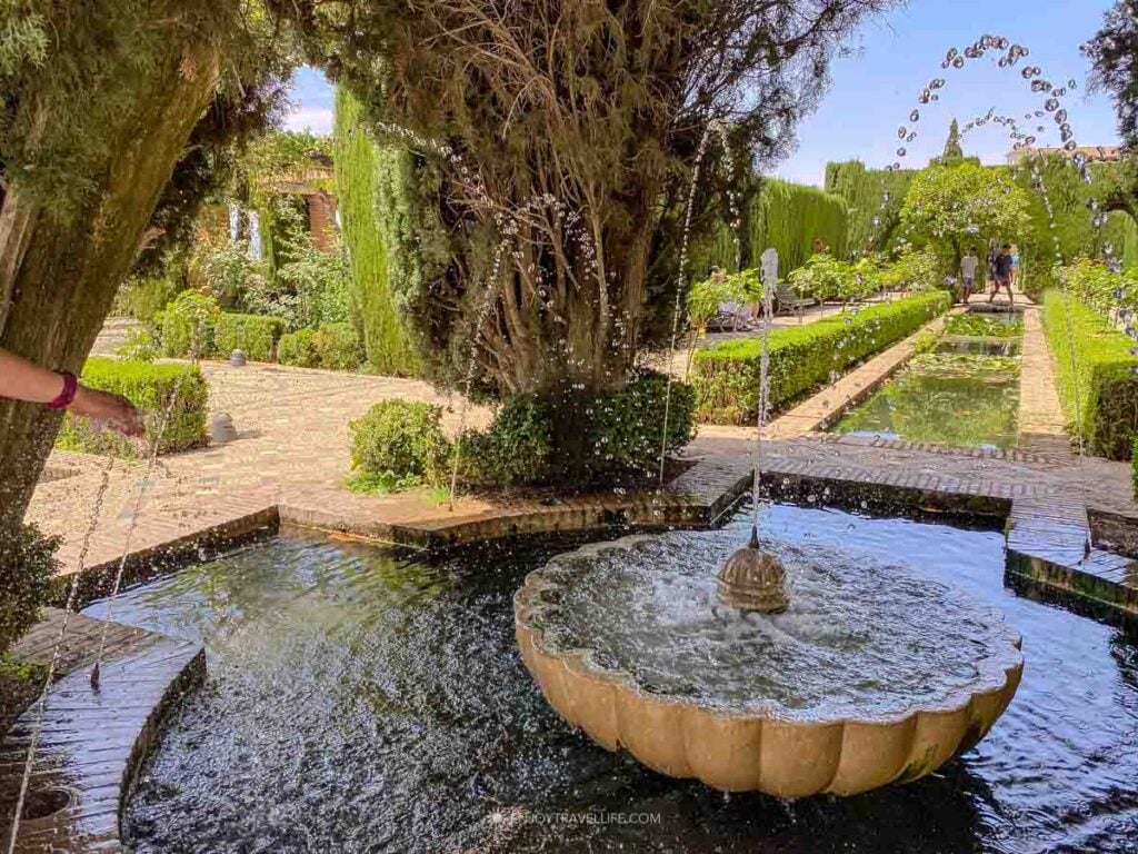 Ornate stone fountain with water jets in the Generalife gardens in Granada, Spain, with cypress trees and clipped hedges lining a walkway behind it.