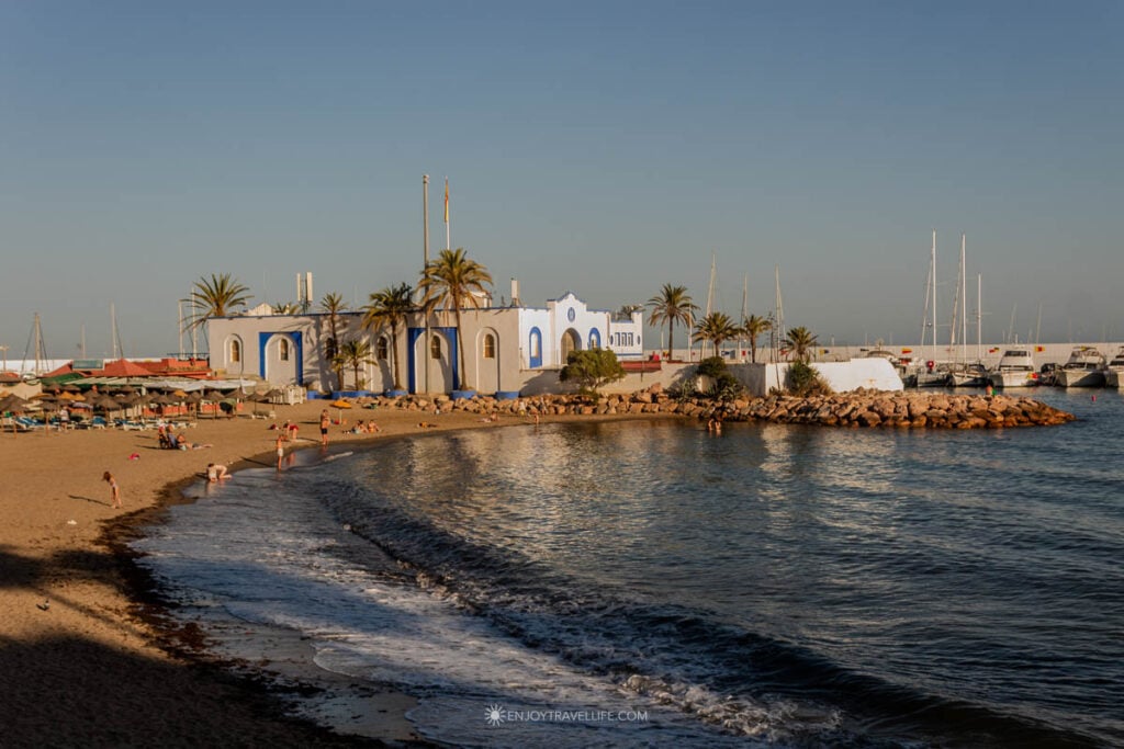 Golden hour on Marbella's beach with a white-and-blue building, palm trees, and marina sailboats in the background.