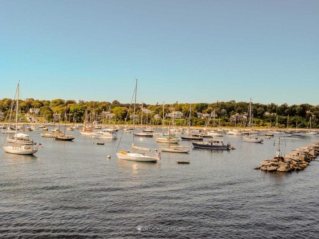 Sailboats moored in Vineyard Haven Harbor with waterfront homes and a stone jetty on a sunny day