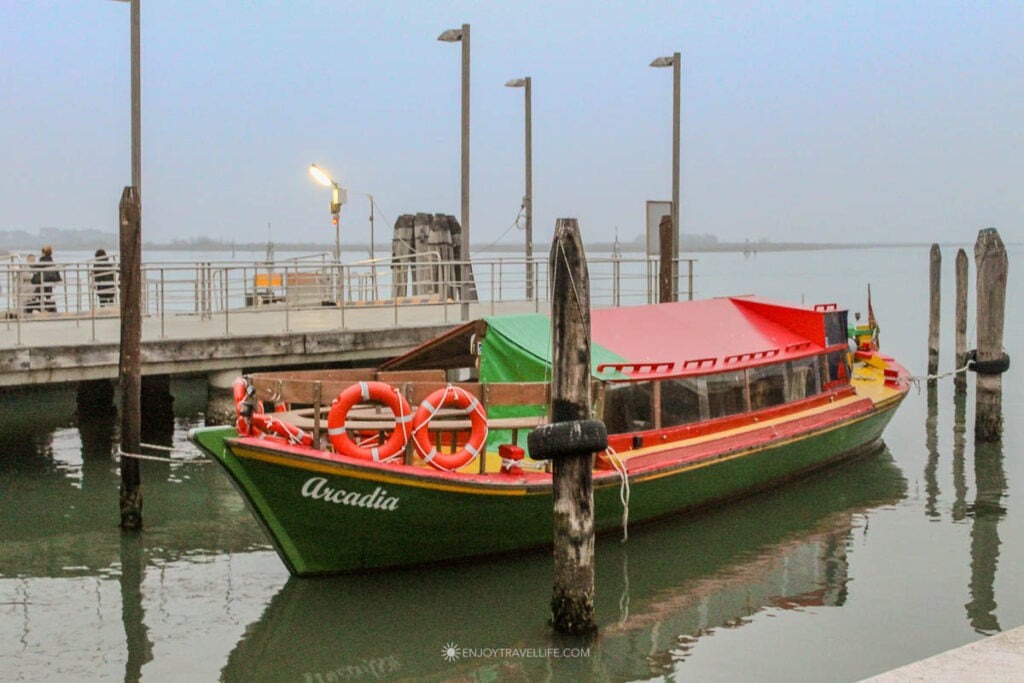 Murano and Burano from Venice: A green and red wooden boat named Arcadia sits moored at a ferry dock in the Venice lagoon on a gray winter day.