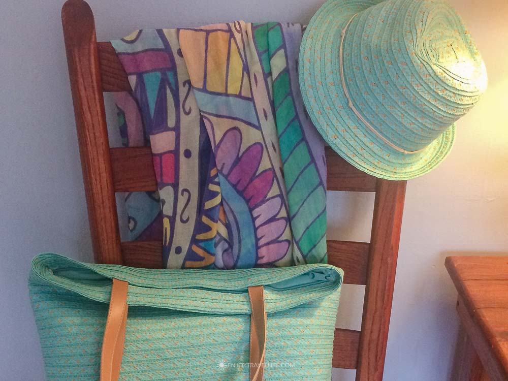 A turquoise straw beach tote and sun hat with a colorful beach towel on a wooden chair