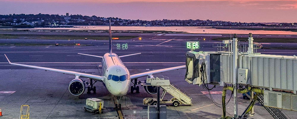 A Delta jet at a Boston Logan gate under a pink and orange sunset sky