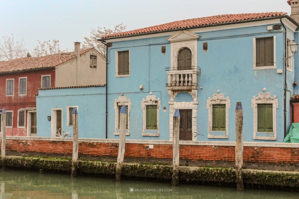 A blue Burano house with an ornate white stone balcony stands on a canal on a gray winter day.