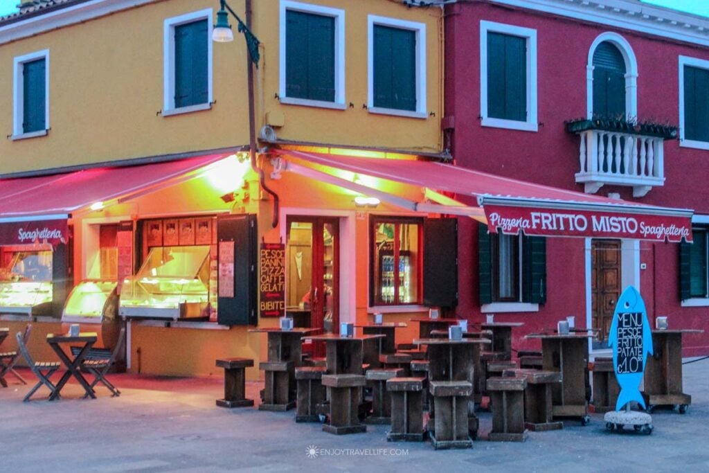 A pink cafe with outdoor tables sits on a Burano canal corner surrounded by colorful houses in winter.