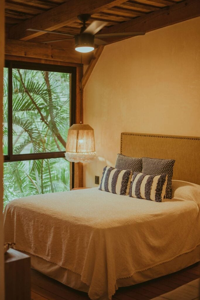 A cozy guesthouse bedroom with a wood-beamed ceiling, a neatly made bed with patterned pillows, and a window looking onto green foliage.
