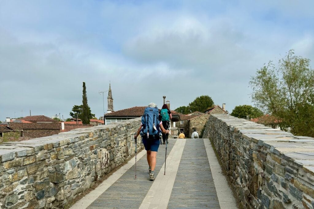 A backpacker with trekking poles walks a stone path toward a village, with other walkers up ahead.