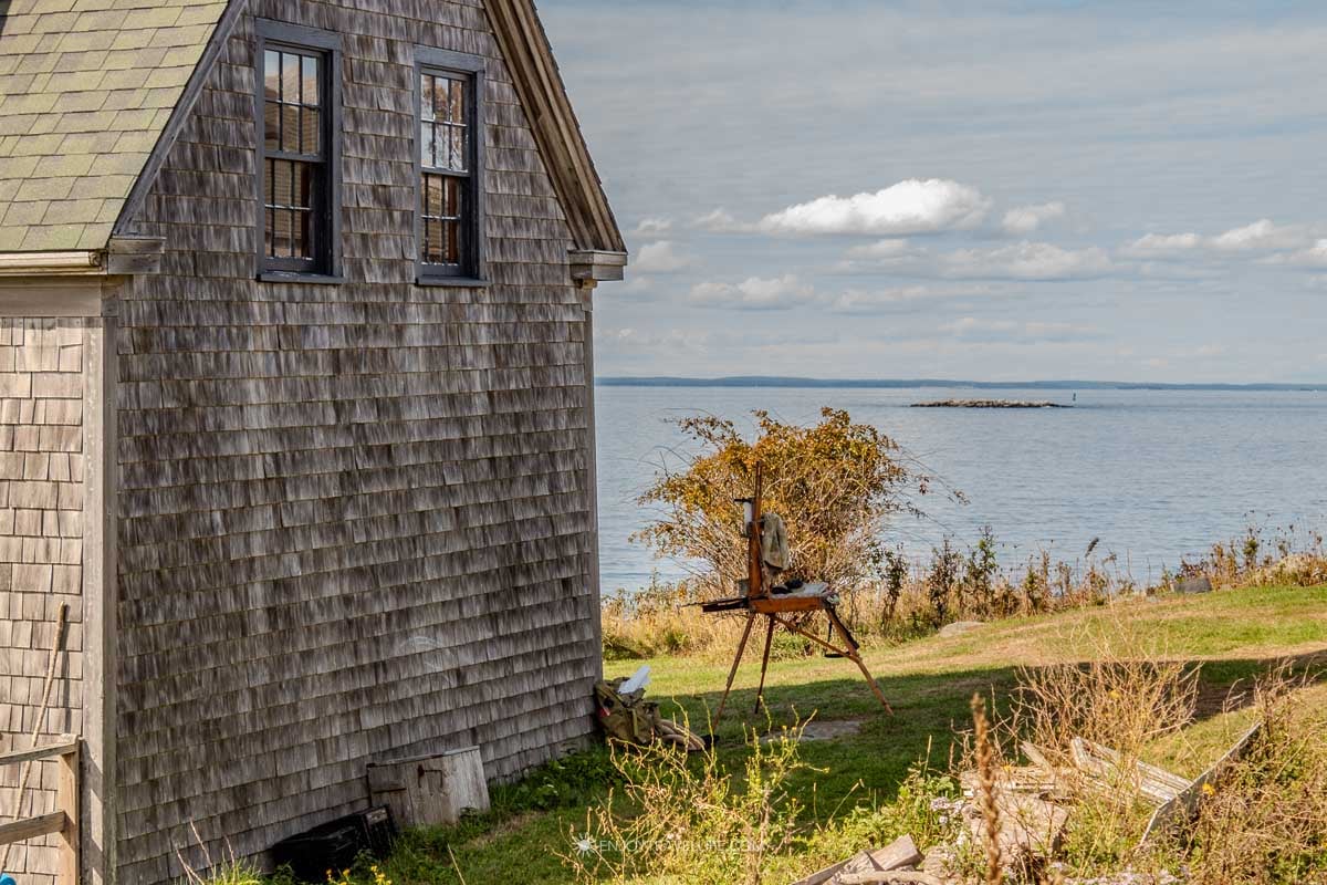 An artist's easel stands on a lawn overlooking a small bay beside a weathered shingle house, with boats at their moorings on Monhegan Island.