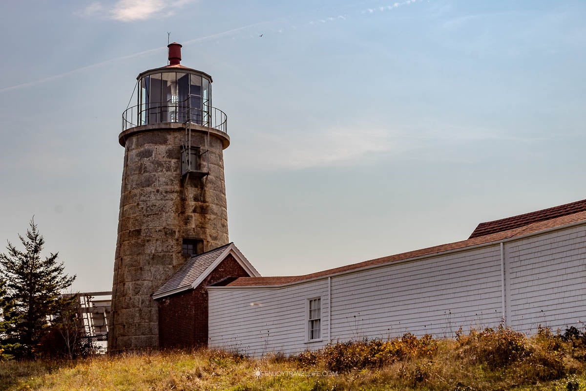 Monhegan Island Light, a white lighthouse tower, rises against a blue sky at the island's highest point.