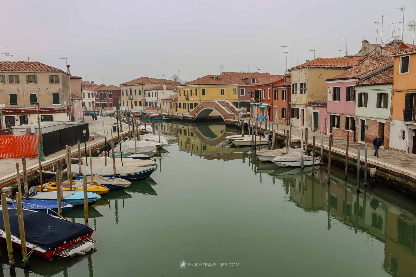 A stone footbridge crosses a quiet Murano canal in winter, with moored boats and colorful houses reflected in the still water.
