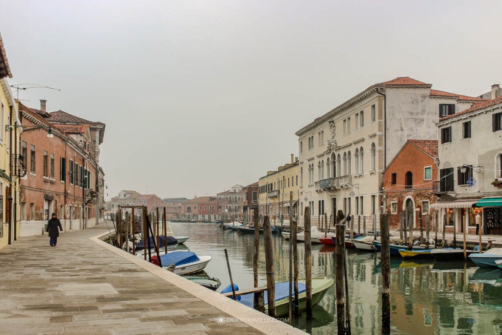 A lone figure walks the stone path beside a quiet Murano canal lined with colorful buildings and moored boats in winter fog.