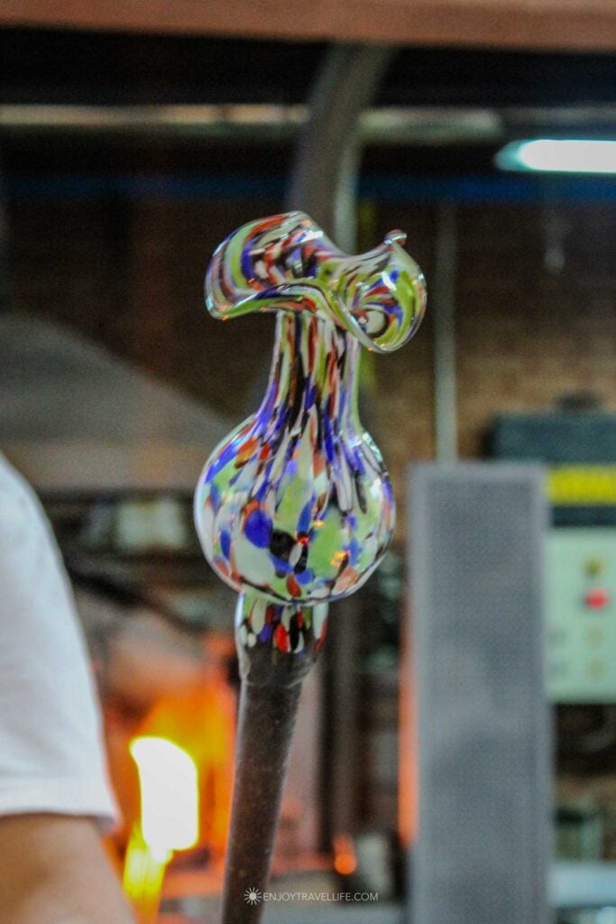 A multicolored glass piece takes shape on the end of a blowpipe as the furnace glows behind it in Murano.