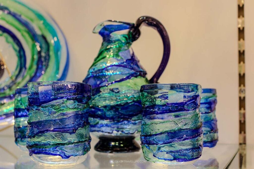 A handblown Murano glass pitcher and matching glasses in swirled blue and green tones sit on display in a showroom.