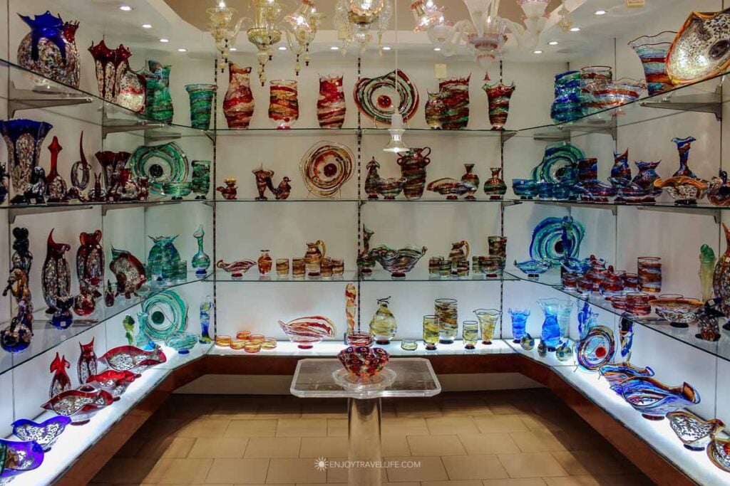Floor-to-ceiling shelves hold colorful Murano glass vases, chandeliers, and sculptures in a bright showroom.