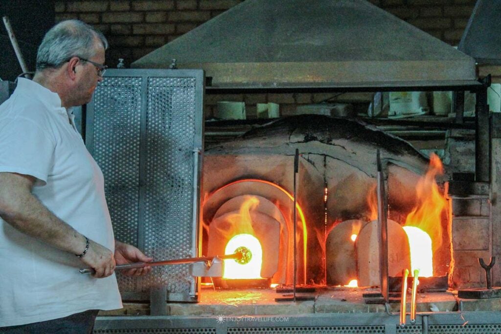 A master glassblower in a white shirt pulls molten glass from a glowing orange furnace during a demonstration in Murano.