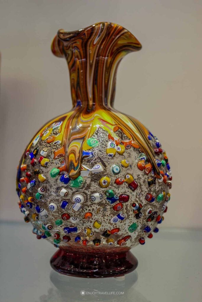 A golden Murano glass vase covered in colorful millefiori dots stands against a cream wall in a showroom.