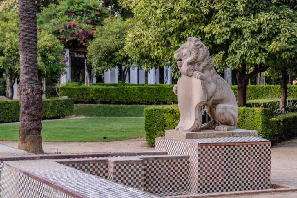 A stone lion statue among clipped hedges and palms in Parque de María Luisa in Seville.