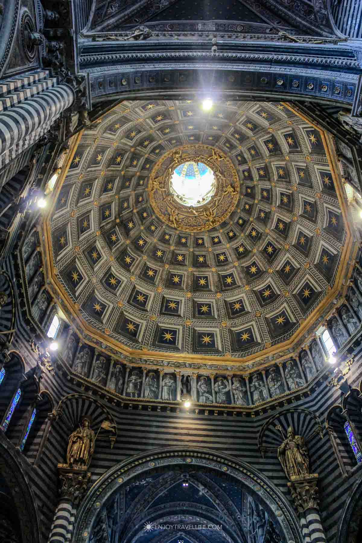 Looking up into the gold-starred coffered dome ceiling inside Siena Cathedral with striped marble columns below