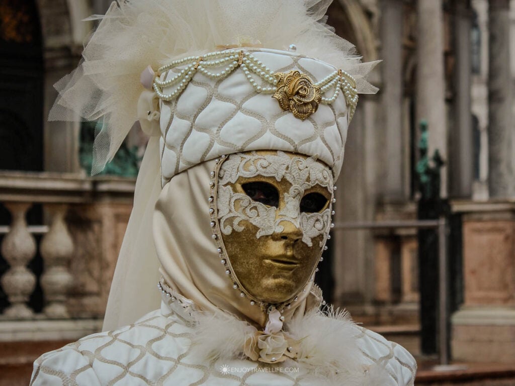 A person in an elaborate white and gold Venice Carnival costume and golden mask poses in front of a basilica in Venice.