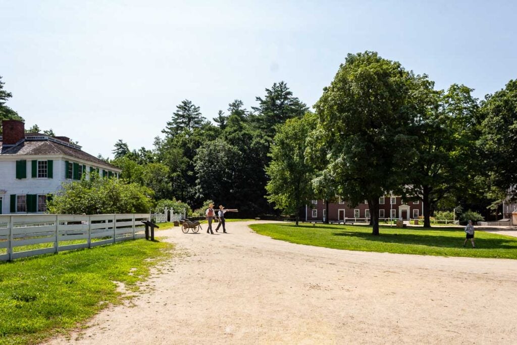 Two costumed historians cross the village common, one carrying boards and the other pulling a wooden handcart.