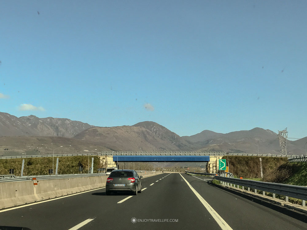 A car heads down an open highway toward hazy mountains under a clear blue sky.