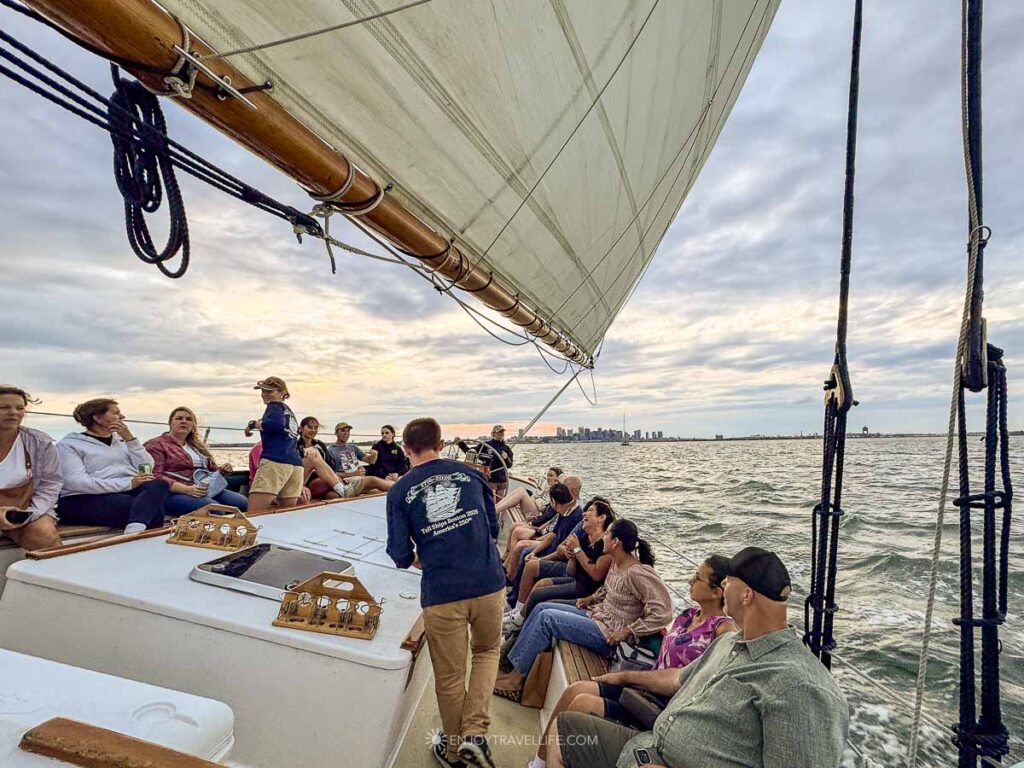 A Boston Harbor sail under full canvas, with guests along the rails and the city skyline low on the horizon.