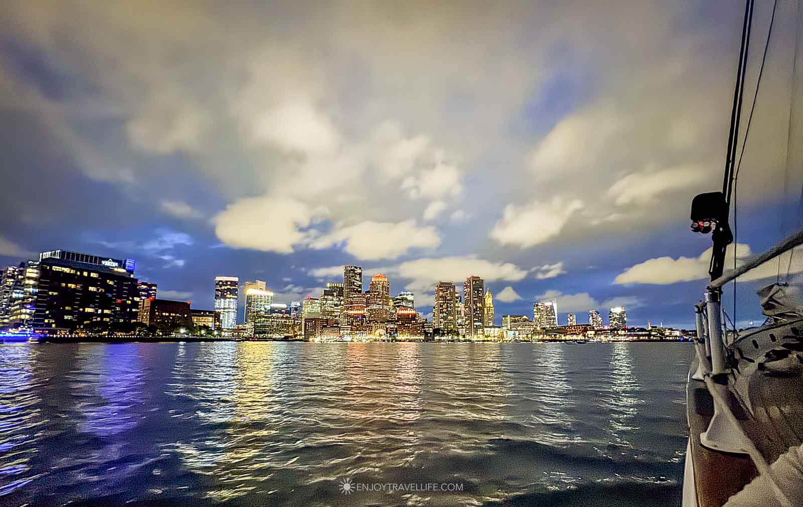 The Boston skyline lit at night under broken cloud, seen across the water from the deck of a schooner.