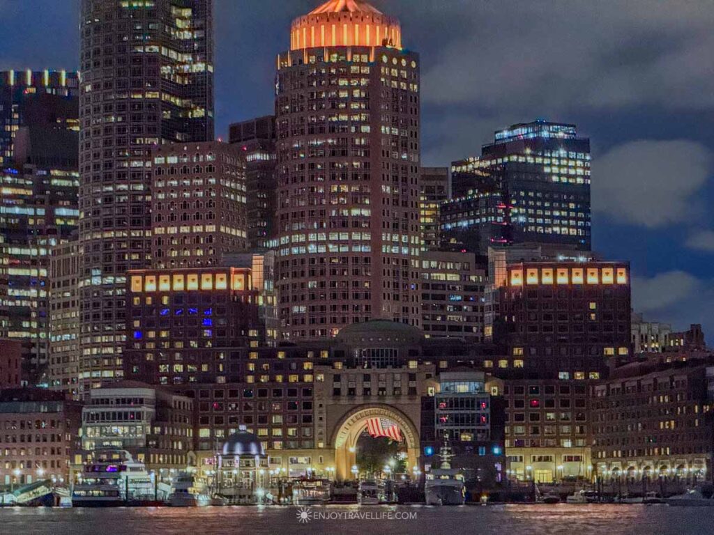 The Boston skyline lit at night with the Rowes Wharf arch glowing below, seen from the water.