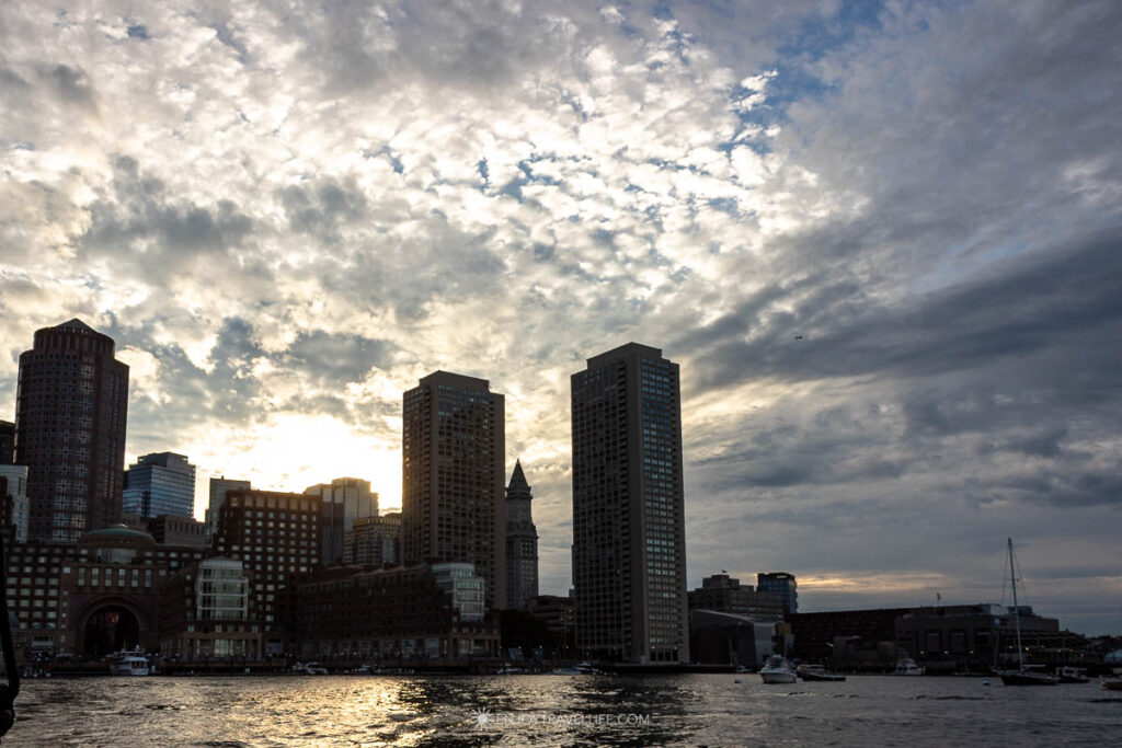 Late sun breaks through cloud above the downtown Boston towers, seen from the middle of the harbor.