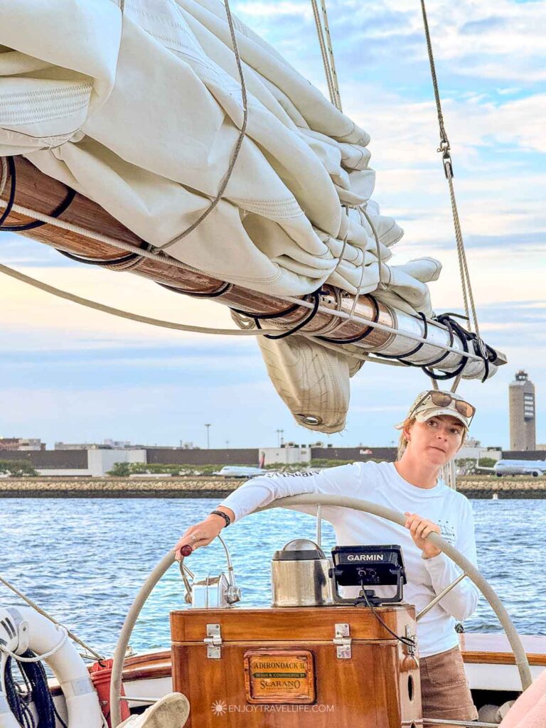 The captain steers the schooner from the wheel in Boston Harbor, with the Logan Airport control tower behind.
