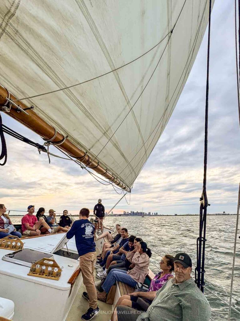 Guests relax along the open deck of a wooden schooner beneath a full sail during an evening in Boston Harbor.