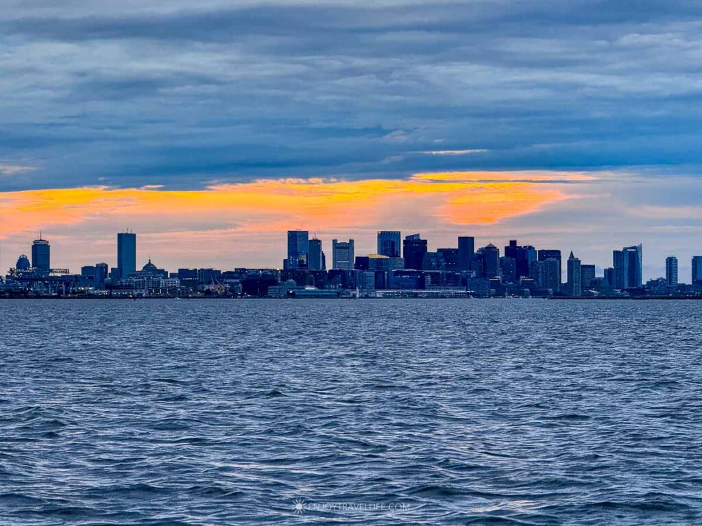 Orange and yellow sunset light spreads behind the Boston skyline, seen from the harbor.