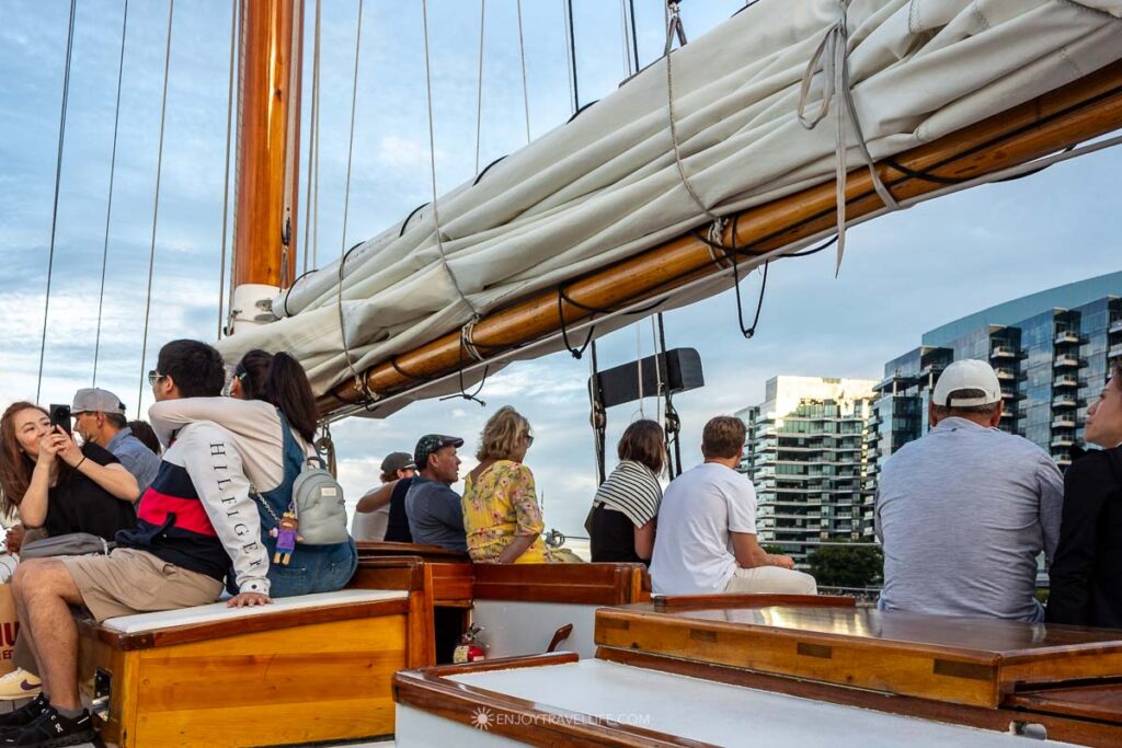 Guests sit along the schooner's varnished rail as the Boston Seaport towers pass behind them.