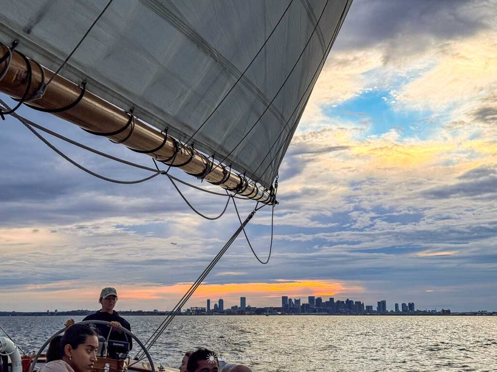 A band of sunset light sits behind the Boston skyline, framed by the schooner's boom and rigging.