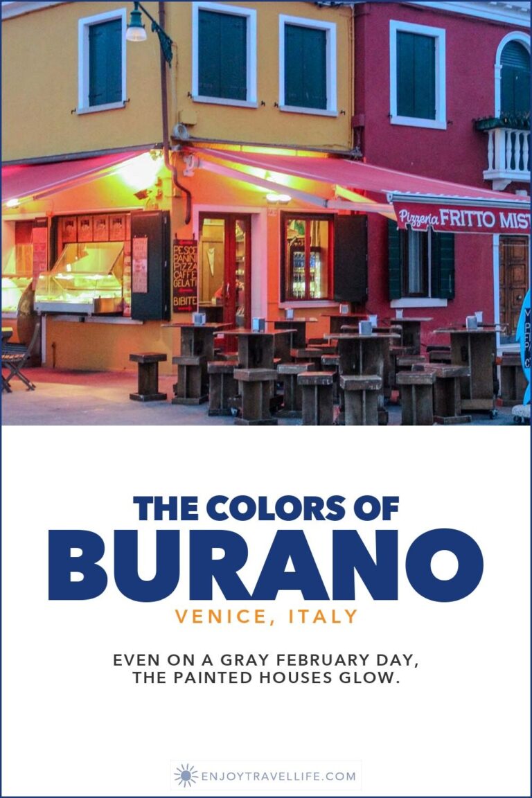 A pink café with outdoor tables on a canal corner in Burano, Italy, beside yellow and red houses.
