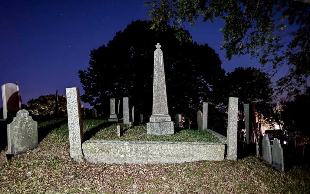 A stone obelisk and headstones on Burial Hill in Plymouth against a deep blue night sky.
