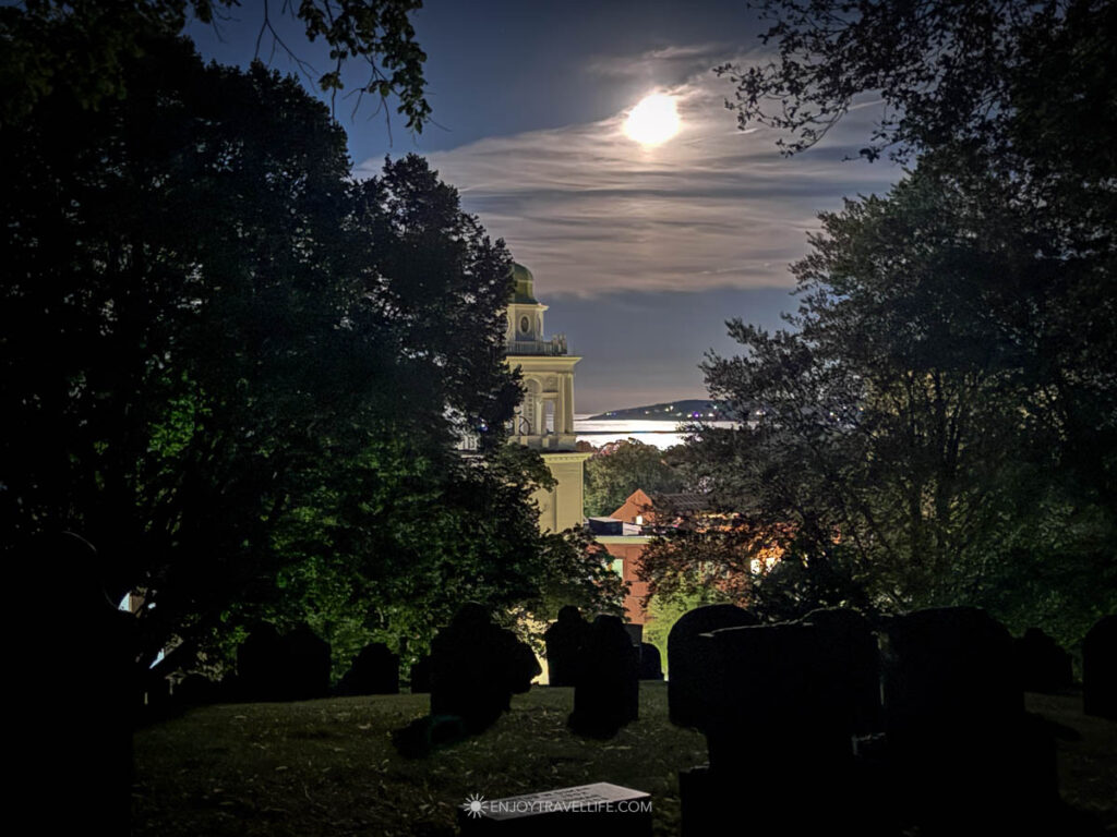 The moon over Plymouth harbor seen from Burial Hill, with headstones silhouetted in the foreground.