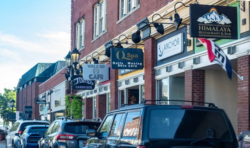 Hanging shop signs and black lanterns line a brick storefront block in downtown Plymouth.