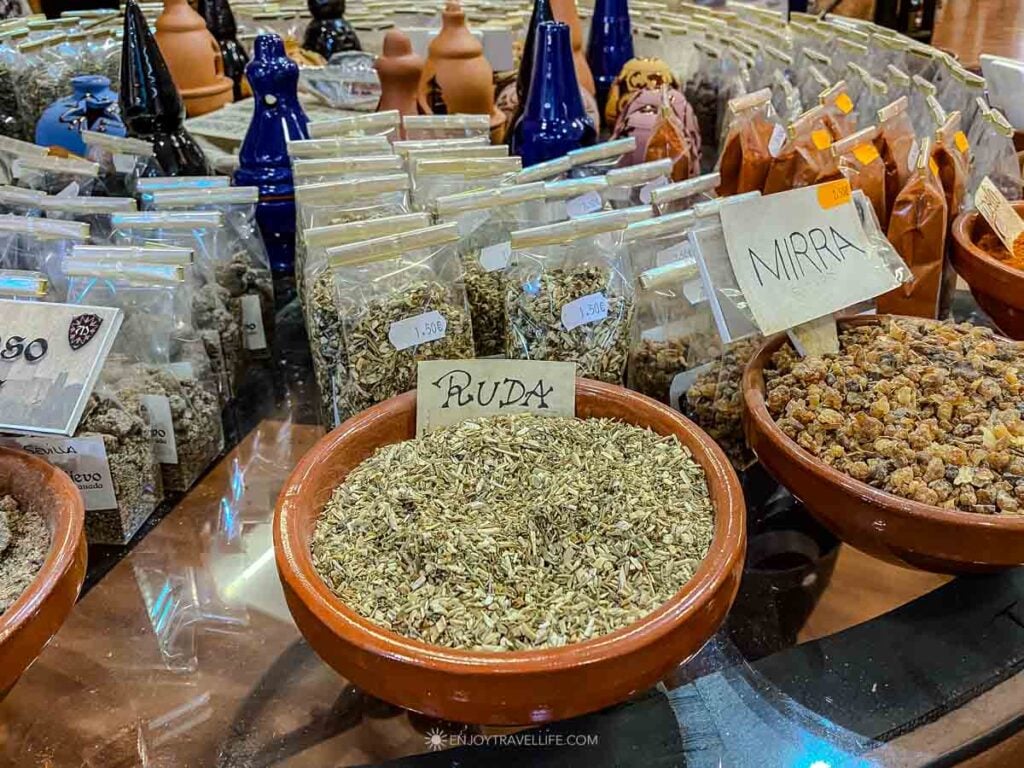 Open sacks of dried herbs and spices with handwritten labels fill a market stall in Granada, Spain.
