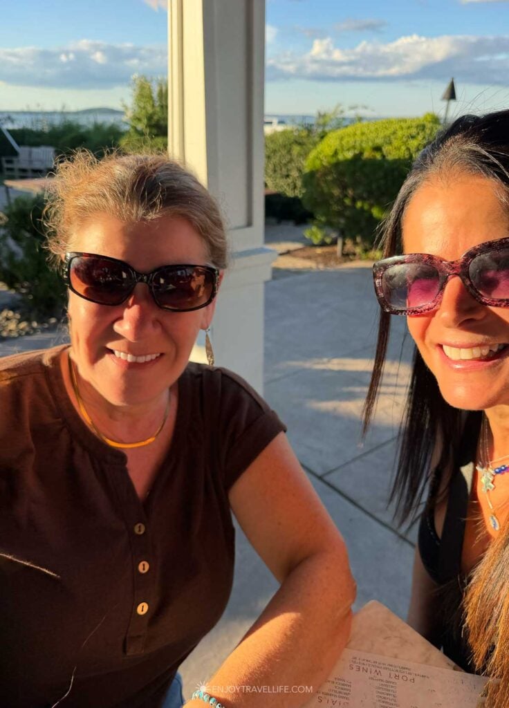 Two women in sunglasses smile together on a restaurant porch overlooking the water in Plymouth.