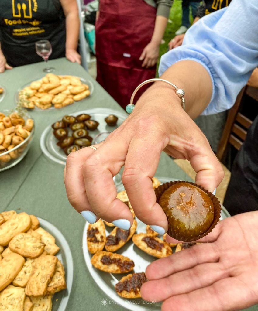 A vendor on a Kalamata food tour in Greece hands a visitor a candied fig, with plates of stuffed dried figs and olive-oil biscuits on the table behind.