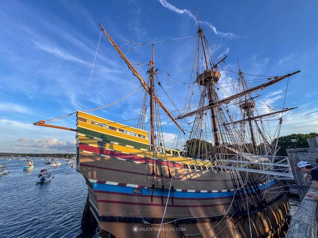 The Mayflower II is docked at a wooden pier in Plymouth Harbor under a blue sky.