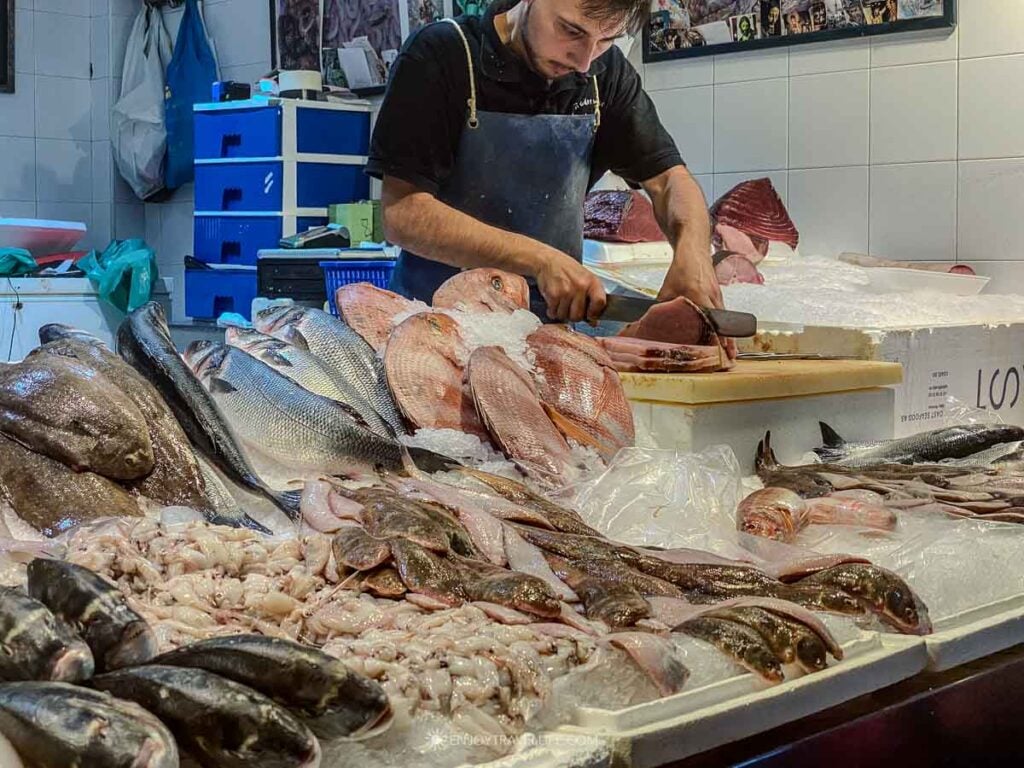 A fishmonger in a blue apron cuts fish to order behind a counter of whole fish on ice at the Mercado de Triana in Seville, Spain.