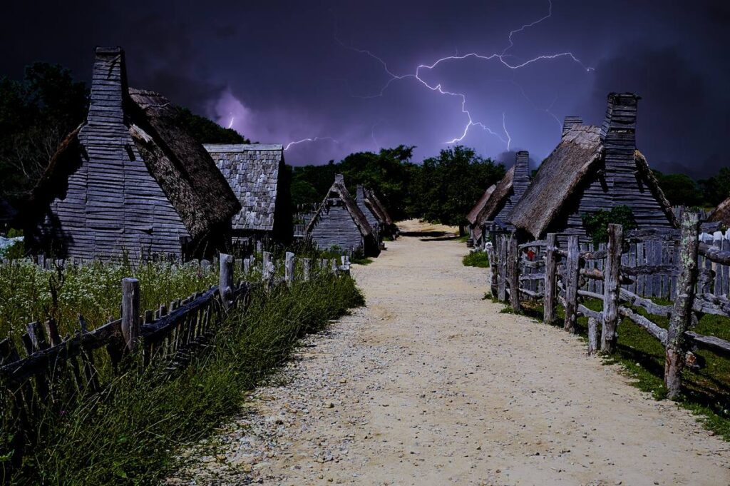 Thatched wooden houses line a gravel lane in the 17th-Century English Village at Plimoth Patuxet Museums under a dark, stormy sky.