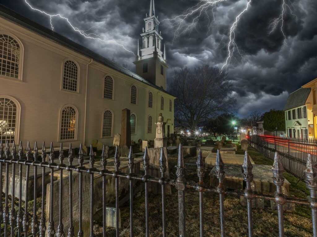 Trinity Church in Newport rises behind an iron fence and its old burying ground under a dramatic night sky.